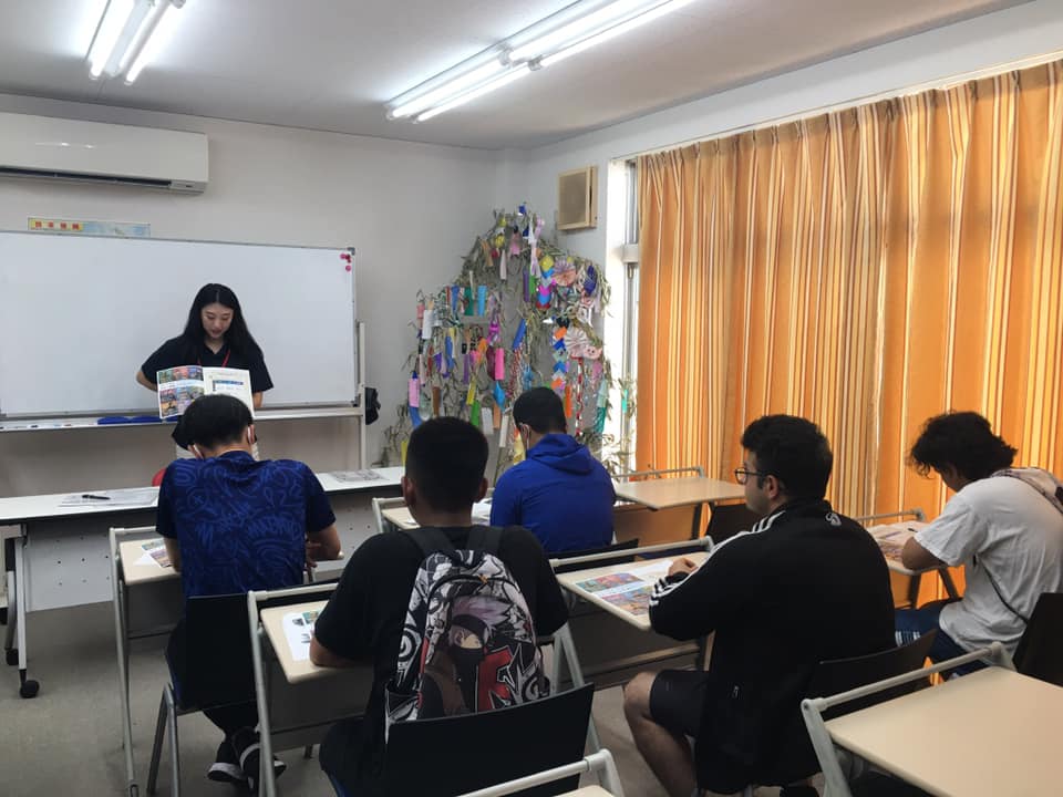 INC日本語学院の授業風景。教室で講師が教材を手に説明し、学生たちが着席して授業を受けている様子。